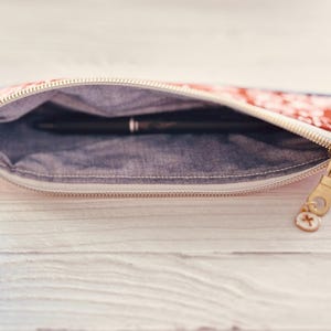 May include: Open pencil case with a floral pattern in red and white, revealing a black pen inside. The case has a denim-like interior and a gold zipper with a charm.