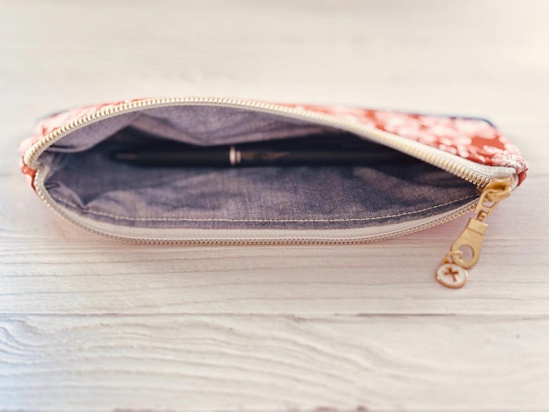 May include: Open pencil case with a floral pattern in red and white, revealing a black pen inside. The case has a denim-like interior and a gold zipper with a charm.