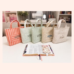 May include: Five fabric tote bags with handles and front pockets, each featuring unique patterns including stripes, floral prints, and gingham checks. The bags display embroidered text like BLESSED and GOD IS GOOD. An open book with a floral cover sits in front.