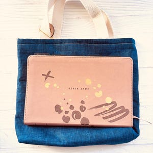 May include: A denim tote bag with a cream-colored handle holds a pink Bible. The Bible cover has gold accents and the words "HOLY BIBLE" printed on it. The tote bag is made of blue denim fabric.