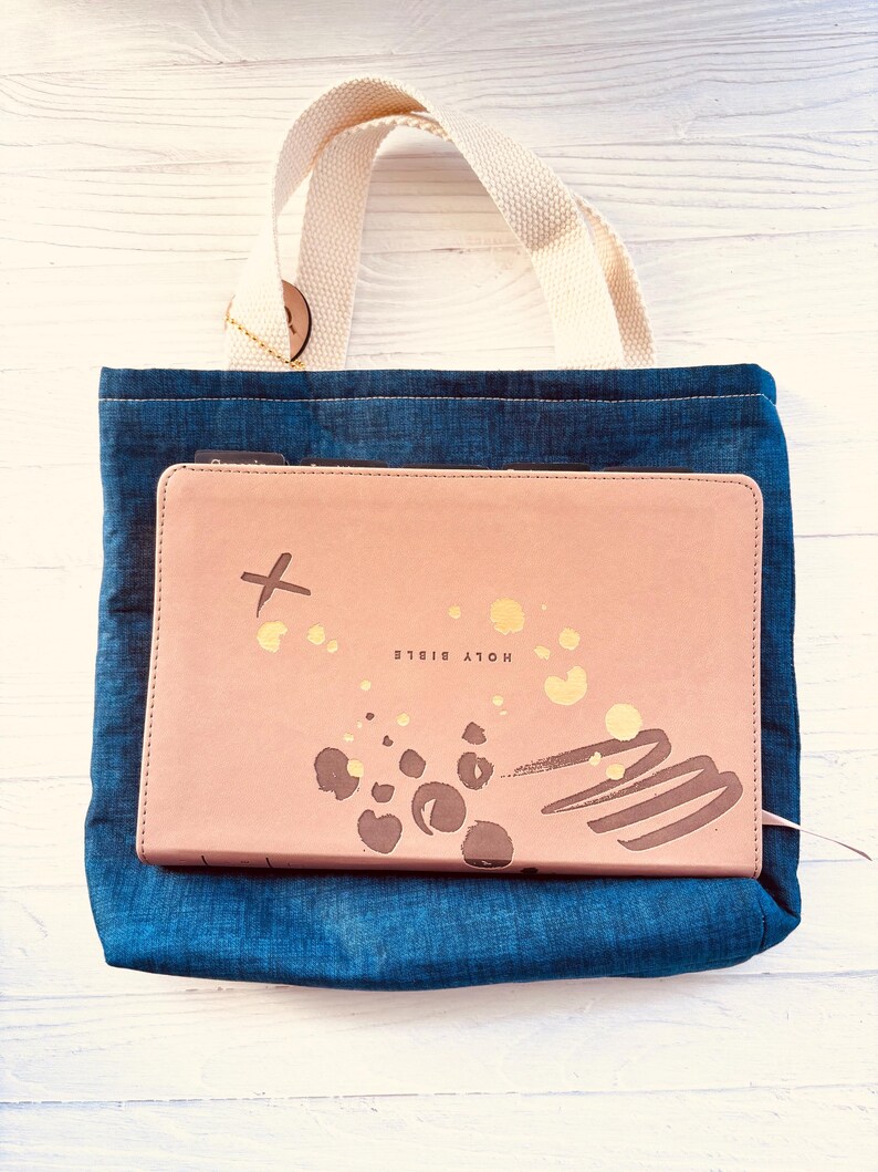 May include: A denim tote bag with a cream-colored handle holds a pink Bible. The Bible cover has gold accents and the words "HOLY BIBLE" printed on it. The tote bag is made of blue denim fabric.