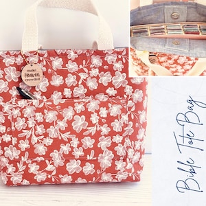 May include: Red floral print tote bag with cream-colored handles and a front pocket. A wooden tag with the words "make Heaven crowded" hangs from the handle. The bag has a denim interior with multiple compartments. The words "Bible Tote Bag" are written on a white background.