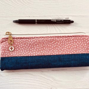 May include: A rectangular pencil case with a pink dotted pattern on top and a dark blue bottom. A black pen rests above the case. The pencil case has a gold zip and a charm with a cross.