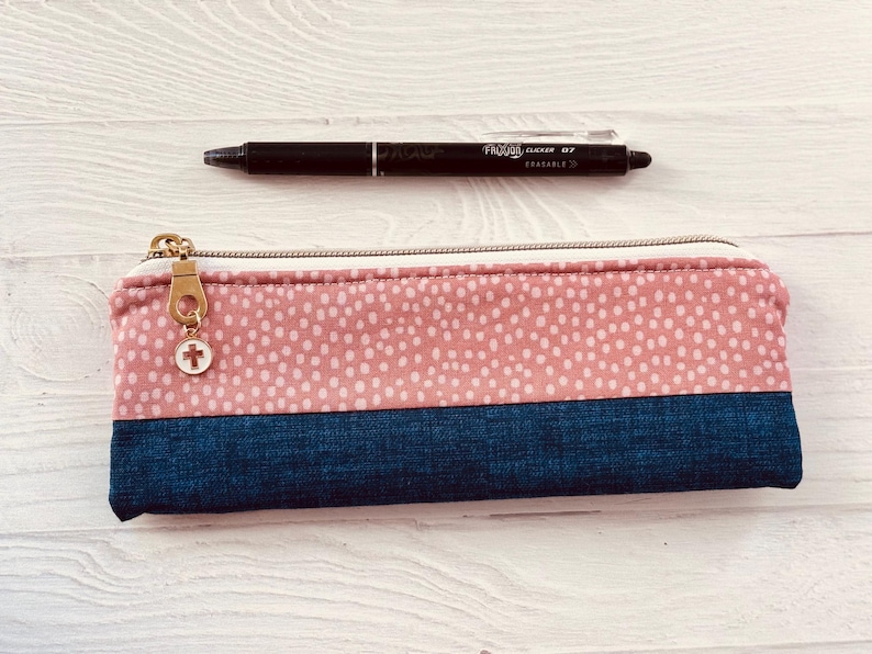 May include: A rectangular pencil case with a pink dotted pattern on top and a dark blue bottom. A black pen rests above the case. The pencil case has a gold zip and a charm with a cross.
