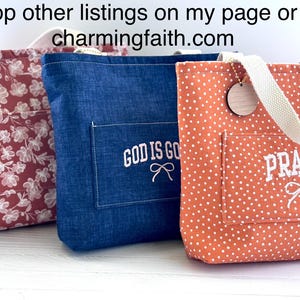May include: Three tote bags in a row. The first is red with white floral patterns, the second is blue with "GOD IS GOOD" embroidered on the pocket, and the third is orange with white polka dots and "PRAYER" embroidered on the pocket. Each has a wooden tag.