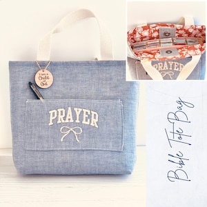 May include: A blue denim tote bag with a white handle and a front pocket. The pocket has the word "PRAYER" embroidered in white, with a bow below. A wooden tag with the text "I am a Child of God" hangs from the handle.