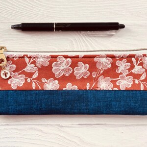 May include: A rectangular pencil case with a floral pattern in white on a rust-colored background, and a dark blue bottom. A black pen rests above the case. The case has a gold zipper and a small charm.
