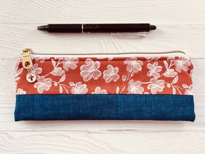May include: A rectangular pencil case with a floral pattern in white on a rust-colored background, and a dark blue bottom. A black pen rests above the case. The case has a gold zipper and a small charm.