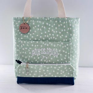 K&ouml;nnte beinhalten: Eine mintgr&uuml;ne Tragetasche mit wei&szlig;en Tupfen, einem cremefarbenen Griff und einem passenden Federm&auml;ppchen. Die Tasche hat eine Vordertasche mit dem gestickten Text "GOD IS GOOD" und ein Holzschild mit der Aufschrift "GIVE IT TO god".
