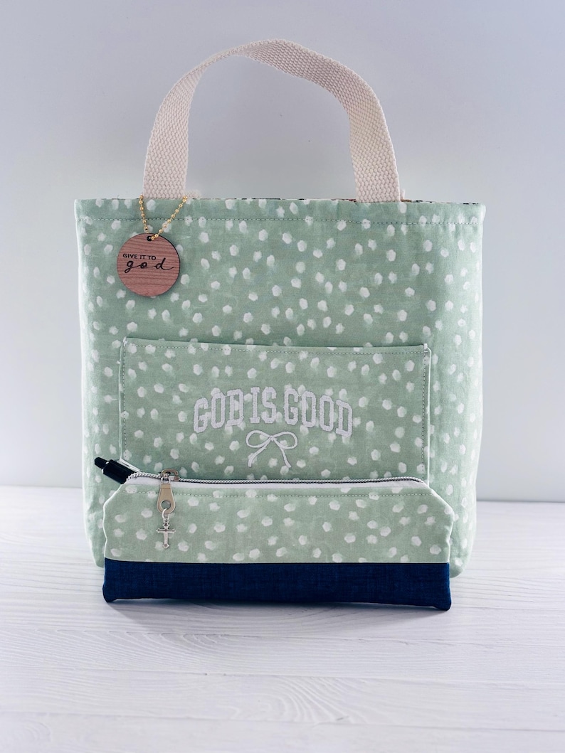 K&ouml;nnte beinhalten: Eine mintgr&uuml;ne Tragetasche mit wei&szlig;en Tupfen, einem cremefarbenen Griff und einem passenden Federm&auml;ppchen. Die Tasche hat eine Vordertasche mit dem gestickten Text "GOD IS GOOD" und ein Holzschild mit der Aufschrift "GIVE IT TO god".