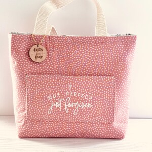 May include: A pink tote bag with a white dotted pattern and a cream-coloured handle. The bag has a front pocket with the text "NOT PERFECT just forgiven". A wooden tag with "faith over fear" hangs from the handle.