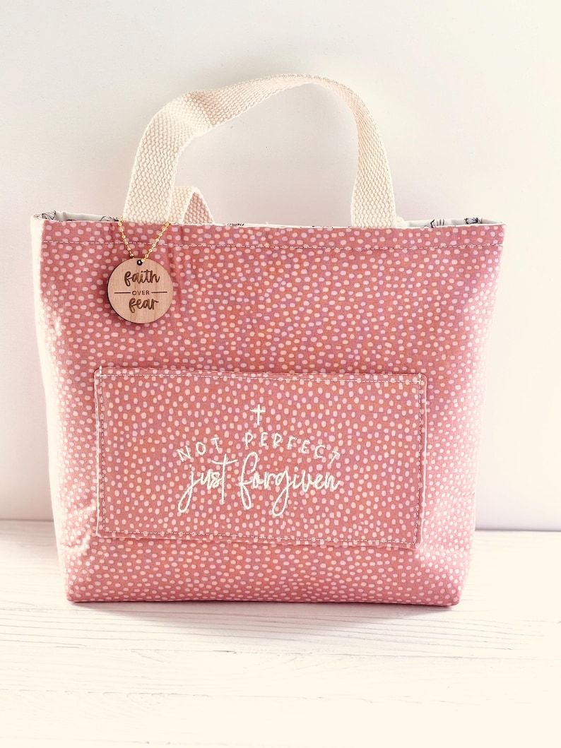May include: A pink tote bag with a white dotted pattern and a cream-coloured handle. The bag has a front pocket with the text "NOT PERFECT just forgiven". A wooden tag with "faith over fear" hangs from the handle.