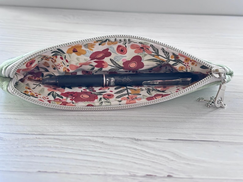 K&ouml;nnte beinhalten: Hellgr&uuml;nes Federm&auml;ppchen mit floralem Innenfutter, ge&ouml;ffnet, um einen schwarzen Stift zu zeigen. Der Innenstoff hat ein Muster aus roten, gelben und wei&szlig;en Blumen mit gr&uuml;nen Bl&auml;ttern. Der Rei&szlig;verschluss ist silberfarben.