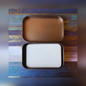 May include: An open, rectangular, bronze-colored tin with a white, rectangular insert. The tin's lid is open, revealing the interior. The background is a wooden surface with blue, gold, and brown tones.
