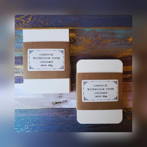 May include: Two packages of Canson XL watercolor paper. Each package is wrapped in brown paper and features a white label with the product details. The labels read "CANSON XL WATERCOLOR PAPER cold pressed 140-lb. 300g".