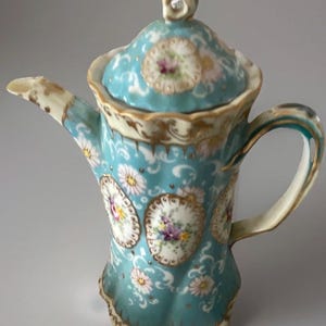 May include: A light blue porcelain teapot with a matching lid, featuring white floral patterns, gold trim, and oval medallions containing colorful painted flower bouquets. The teapot has a curved handle, a scalloped rim, and a pointed spout.