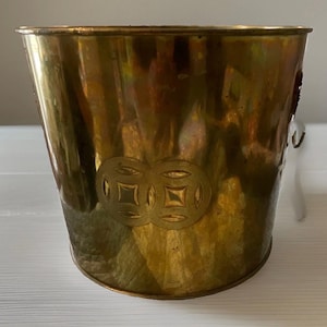 Puede incluir: Un cubo cilíndrico de latón con un acabado metálico pulido. El exterior presenta un diseño grabado de dos monedas circulares entrelazadas. El recipiente tiene una abertura ancha en la parte superior y una pequeña asa de metal en el lateral.