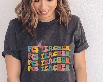 May include: A dark grey heather t-shirt with the text "FCS TEACHER" repeated four times in rainbow colors. A small butterfly graphic is above the text.