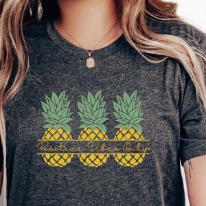 May include: A dark grey heather t-shirt with a graphic of three yellow pineapples with green tops. The text "Positive Vibes Only" is printed in a script font above the pineapples.