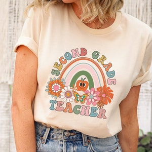 May include: A cream colored t-shirt with a retro rainbow design and the text "Second Grade Teacher" in colorful letters. The rainbow has a smiling apple face in the center.