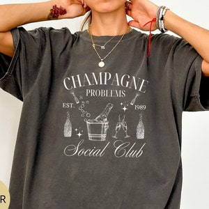 May include: A dark gray t-shirt with a white graphic design that says "Champagne Problems Social Club" with a champagne bottle and glasses in a bucket of ice. The text also says "Est. 1989".