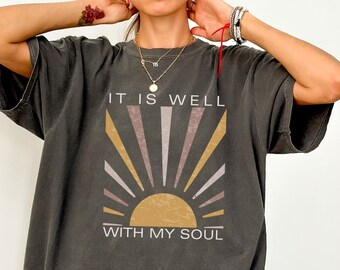 May include: A dark gray oversized t-shirt with a graphic print of a sunrise and the text "It is well with my soul".