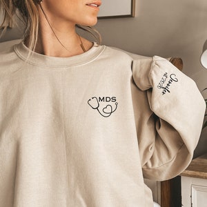May include: A beige sweatshirt with a stethoscope and heart graphic on the front and the text "Jennifer 07.2020" on the sleeve.