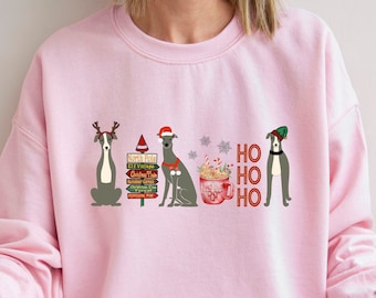 May include: A light pink sweatshirt with a Christmas-themed design featuring three greyhounds wearing Santa hats and reindeer antlers. The design also includes a mug of hot chocolate with marshmallows and the text "HO HO HO".