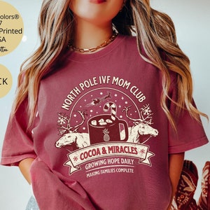 Puede incluir: Una camiseta roja con un dise&ntilde;o gr&aacute;fico blanco que dice "North Pole IVF Mom Club" con una taza de chocolate caliente y renos. El texto debajo del gr&aacute;fico dice "Cocoa & Miracles", "Growing Hope Daily", y "Making Families Complete".