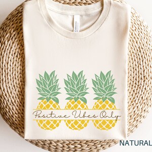 May include: A beige t-shirt featuring a graphic of three yellow pineapples with green tops. The pineapples are arranged in a row with the text "Positive Vibes Only" written in a cursive font below them.