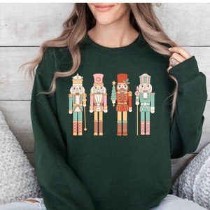 May include: A dark green sweatshirt featuring a colorful graphic of four nutcrackers. Each nutcracker is uniquely designed with different colors and details, including crowns, hats, and holding various items. The sweatshirt is labeled "Forrest Green".