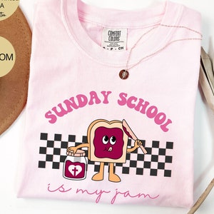 May include: Pink t-shirt with a graphic of a slice of bread with jelly and a cross on a jar. The text "SUNDAY SCHOOL is my jam" is printed on the shirt.