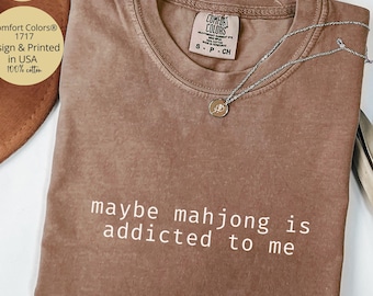 May include: A brown t-shirt with the text "maybe mahjong is addicted to me" printed in white on the front. The shirt is made of 100% cotton.