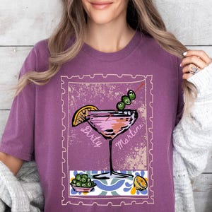 K&ouml;nnte beinhalten: Ein lila T-Shirt mit einer Grafik eines Martini-Cocktails, Oliven und einer Zitronenscheibe. Die Worte "Dirty Martini" sind auf dem Bild aufgedruckt. Das Shirt hat eine einfarbige lila Farbe.
