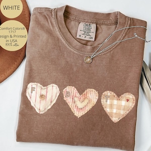 May include: A brown Comfort Colors t-shirt with three heart appliqués spelling "FDT". The hearts are in various patterns and colors, with a small button and a tiny red heart. A silver necklace with a pendant rests on the shirt.