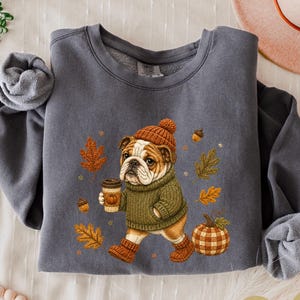 May include: A dark grey crewneck sweatshirt featuring a graphic of a bulldog wearing a brown knit hat, a green jumper, and brown boots while holding a coffee cup. The design includes scattered autumn leaves, acorns, and a plaid pumpkin.