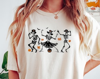 Op de afbeelding: Ivoor kleurig T-shirt met een zwarte afbeelding van drie dansende skeletten. De skeletten dragen hoeden en een rok, met vleermuizen, bladeren en pompoenen. Het shirt bevat de tekst "Comfort Colors 1717 Design & Printed in USA 100% cotton."