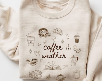 May include: Sand-colored sweatshirt with a crew neck, featuring a coffee-themed graphic. The design includes coffee cups, a croissant, coffee beans, and the words "coffee weather" in a handwritten style. The sweatshirt is folded.