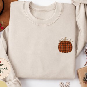 May include: A sand-colored crewneck sweatshirt featuring a small embroidered pumpkin on the left chest. The pumpkin has a brown and black plaid pattern with a tan stem and two small bows. A circular tag reads Faux Patchwork Print and Sand.