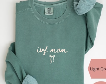 May include: Light green sweatshirt with the text "ivf mom" in cursive script and a bow graphic. The sweatshirt is made of super soft material and is printed in the USA. The color is light green.