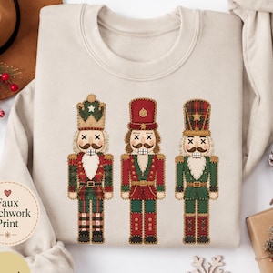 May include: A sand-coloured crewneck sweatshirt featuring a graphic print of three nutcracker figures. Each nutcracker is designed with a faux patchwork aesthetic in shades of red, green, and gold, displaying stitched details and X-shaped eyes.