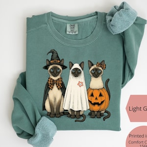 May include: Light green sweatshirt featuring three cartoon cats dressed in Halloween costumes. One cat wears a witch hat and cape, another is in a ghost costume, and the third is in a pumpkin. The sweatshirt is printed in the USA and is super soft.
