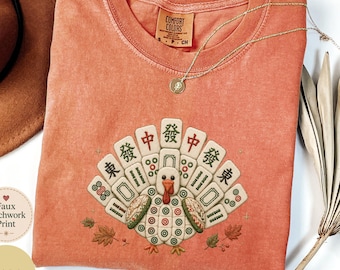 May include: A terracotta-colored short-sleeve t-shirt featuring a graphic of a turkey constructed from mahjong tiles. The turkey's tail feathers are arranged in a fan shape using various mahjong tiles with Chinese characters and green circular patterns.