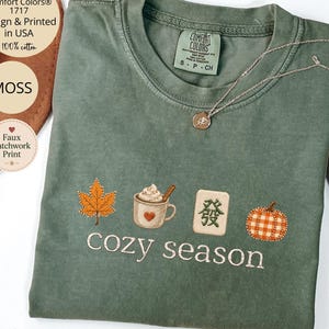 May include: A moss green t-shirt featuring an embroidered design of an orange maple leaf, a mug of hot cocoa with a heart, a mahjong tile with a green character, and a plaid pumpkin, with the text cozy season printed below the graphics.