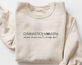 Puede incluir: Una sudadera beige con el texto "Gymnastics Mom Era where chaos meets chalk dust" impreso en la parte delantera.