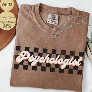 May include: A brown Comfort Colors t-shirt with the word "Psychologist" in a retro font over a black and white checkered pattern. A silver necklace with a pendant rests on the shirt. The shirt is labeled "Design & Printed in USA 100% cotton."