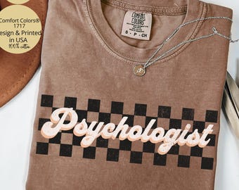 May include: A brown Comfort Colors t-shirt with the word "Psychologist" in a retro font over a black and white checkered pattern. A silver necklace with a pendant rests on the shirt. The shirt is labeled "Design & Printed in USA 100% cotton."