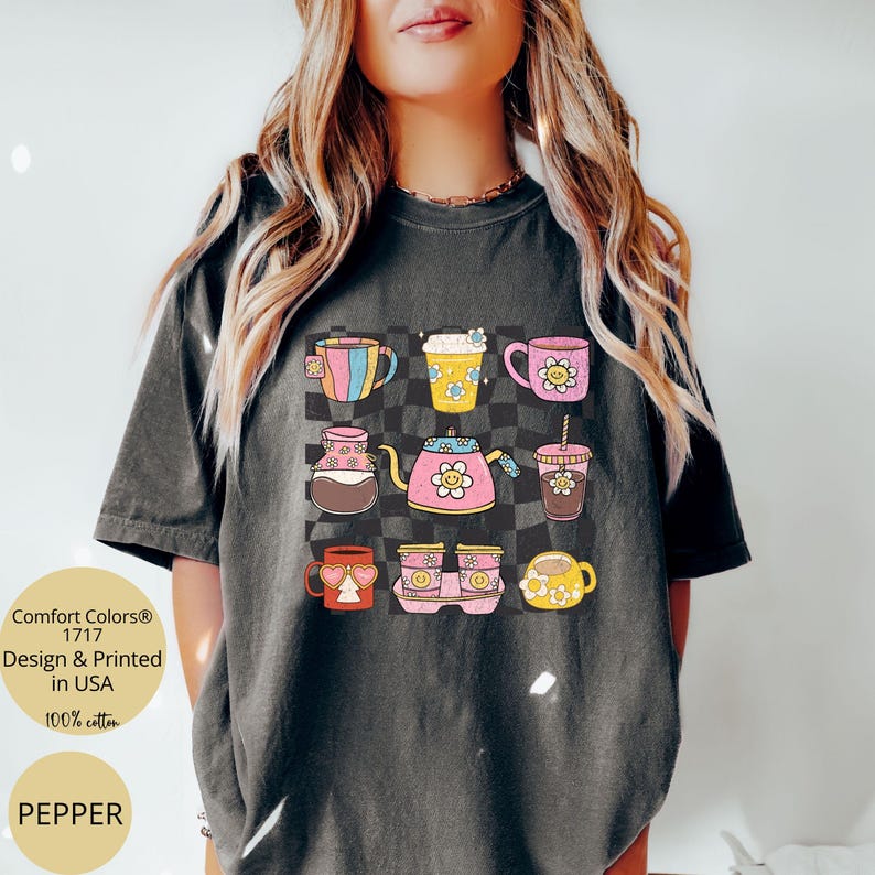 May include: A dark grey t-shirt featuring a graphic of colourful mugs and coffee cups with smiley faces. The text "Comfort Colors&reg; 1717 Design & Printed in USA 100% cotton" is printed on the bottom left corner of the shirt. The word "PEPPER" is printed on the bottom left corner of the shirt.