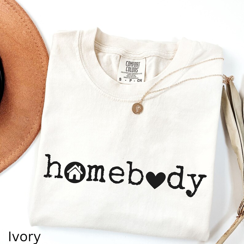 Pode incluir: Camiseta de cor marfim com um gr&aacute;fico preto que diz "homebody" com um &iacute;cone de casa e um cora&ccedil;&atilde;o.
