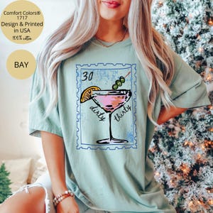 May include: A light green t-shirt featuring a cocktail design with the text "30 dirty thirty". The design includes a pink cocktail with an olive garnish and a lemon slice. The shirt is made of 100% cotton.